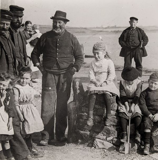 Black and white photo with four men standing in warm, plain clothing, some with beards and smoking pipes. Several children sit beside them, and a woman with a baby stands in the background.
